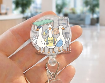 May include: A clear, round badge reel featuring three cartoon geese in a rehab setting. One goose wears a headband, another a "REHAB TEAM" shirt, and the third holds crutches. The background includes a massage table, weights, and a box.