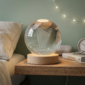 May include: A clear glass globe lamp with a harp design etched on the surface. The lamp sits on a round wooden base and is illuminated from within. The name "Samantha" is written in script below the harp.