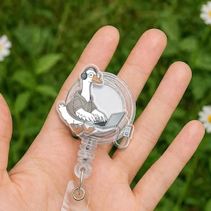 May include: A clear retractable badge reel featuring a cartoon goose wearing headphones and a gray hoodie, working on a laptop. The goose is white with an orange beak and feet. The reel has a silver clip and a clear plastic strap.