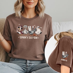 May include: Brown t-shirt featuring a Halloween-themed graphic of geese dressed in costumes, including a witch, mummy, and ghost. The text "Spooky See-Son" is below the geese. A smaller graphic is on the sleeve with the name "Jennifer".