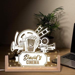 May include: A lighted cinema-themed sign with a wooden base. The sign features a film reel, popcorn, a megaphone, and a movie camera. The text "David's CINEMA" is displayed on the sign. The sign is on a wooden desk with a laptop and a plant.
