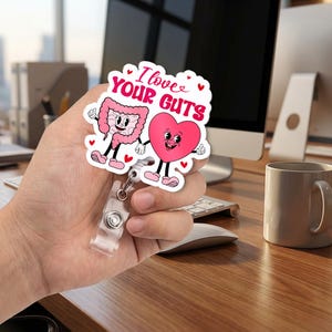 May include: A white badge reel with a cartoon illustration of a pink heart and a pink intestine holding hands. The text "I love your guts" is written in pink and red. The badge reel has a clear plastic strap and a metal clip.