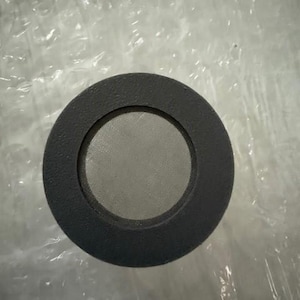 May include: A circular, dark grey metal object with a mesh screen centre. The object has a thick, rounded edge and a fine mesh interior. The item is likely a filter or a component of a larger device.