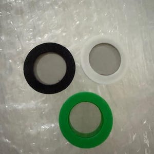 May include: Three circular rubber gaskets in black, white, and green. Each gasket has a hollow centre, creating a ring shape. The gaskets are arranged on a crinkled, translucent surface.