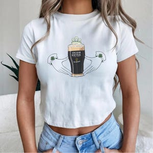 May include: White t-shirt featuring a graphic of a pint of Guinness beer labeled "Irish Extra" held by two hands with shamrock accents. The beer has a frothy head and is topped with a crown. The shirt is a cropped style.