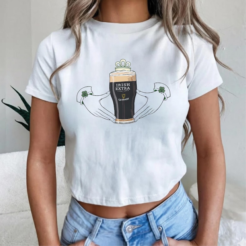 May include: White t-shirt featuring a graphic of a pint of Guinness beer labeled "Irish Extra" held by two hands with shamrock accents. The beer has a frothy head and is topped with a crown. The shirt is a cropped style.