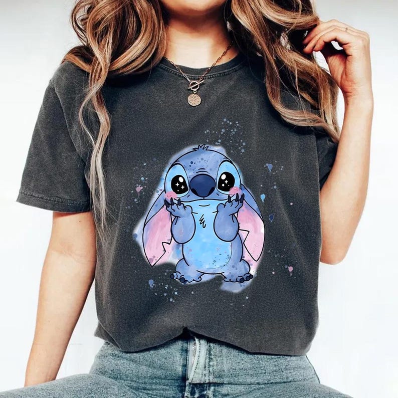 Puede incluir: Camiseta gris oscuro con una ilustraci&oacute;n de dibujos animados de Stitch, un personaje alien&iacute;gena azul con ojos grandes y orejas rosas. El gr&aacute;fico est&aacute; rodeado de salpicaduras de pintura azules y rosas. Prenda de vestir informal.