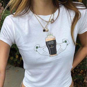 May include: White t-shirt featuring a graphic of a pint of Guinness beer labeled "Irish Extra" held by two hands with shamrock accents. The design is centered on the shirt, with a simple, clean aesthetic. The shirt is short-sleeved.