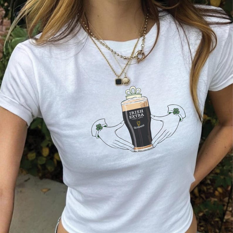 May include: White t-shirt featuring a graphic of a pint of Guinness beer labeled "Irish Extra" held by two hands with shamrock accents. The design is centered on the shirt, with a simple, clean aesthetic. The shirt is short-sleeved.