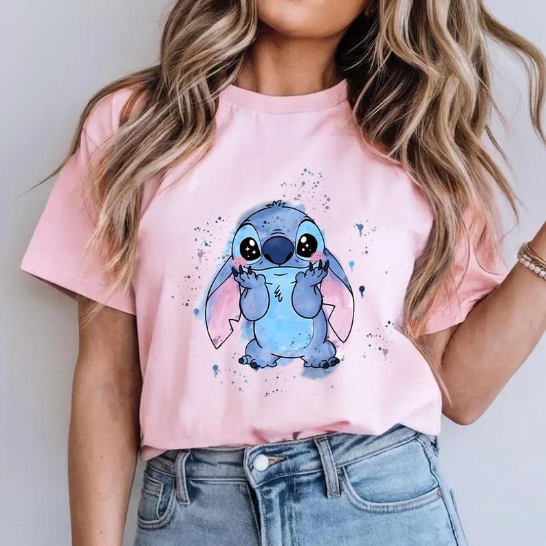 Puede incluir: Camiseta rosa claro con un personaje de dibujos animados azul con ojos y orejas grandes. El personaje est&aacute; rodeado de salpicaduras de pintura azules y moradas. La camiseta es de material suave.