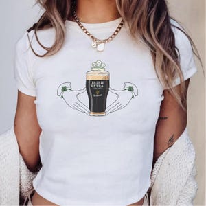 May include: White t-shirt featuring a graphic of a pint of Guinness with the text "IRISH EXTRA". The design includes hands holding the pint and shamrocks. A gold chain necklace is visible.