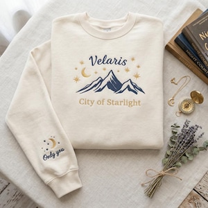 May include: Cream-colored sweatshirt with embroidered design. The design features the text "Velaris City of Starlight" above a mountain range, a crescent moon, and stars. The left sleeve has a moon and stars design with the text "Only you".