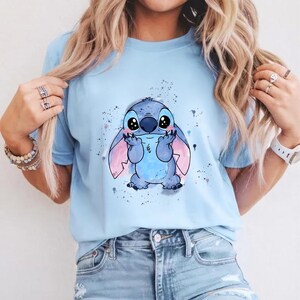 Puede incluir: Camiseta azul claro con una ilustraci&oacute;n de dibujos animados de Stitch de Lilo & Stitch. El personaje es azul con grandes ojos negros y orejas rosas, sobre un fondo de salpicaduras de pintura. La camiseta es de material suave.