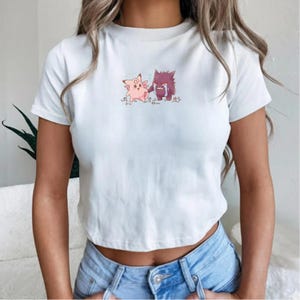 May include: White crop top featuring a cartoon illustration of two characters. One is pink with a smiling expression, and the other is purple and crying. The design is centered on the chest.