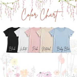 May include: A color chart displays five short-sleeved t-shirts in black, white, pink, natural, and baby blue. The text "Color Chart" is written in cursive script above the shirts. Floral decorations are in the background.