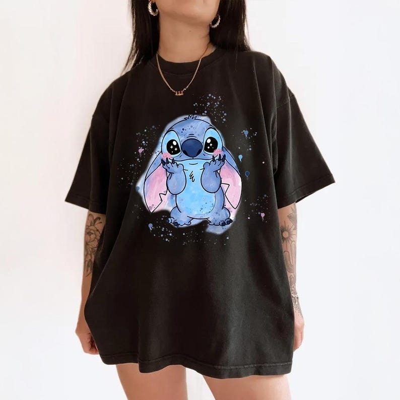 Puede incluir: Camiseta negra con un personaje de dibujos animados azul, con orejas grandes y una expresi&oacute;n amigable. El personaje est&aacute; rodeado de salpicaduras de pintura azul y rosa. La camiseta es de estilo casual.