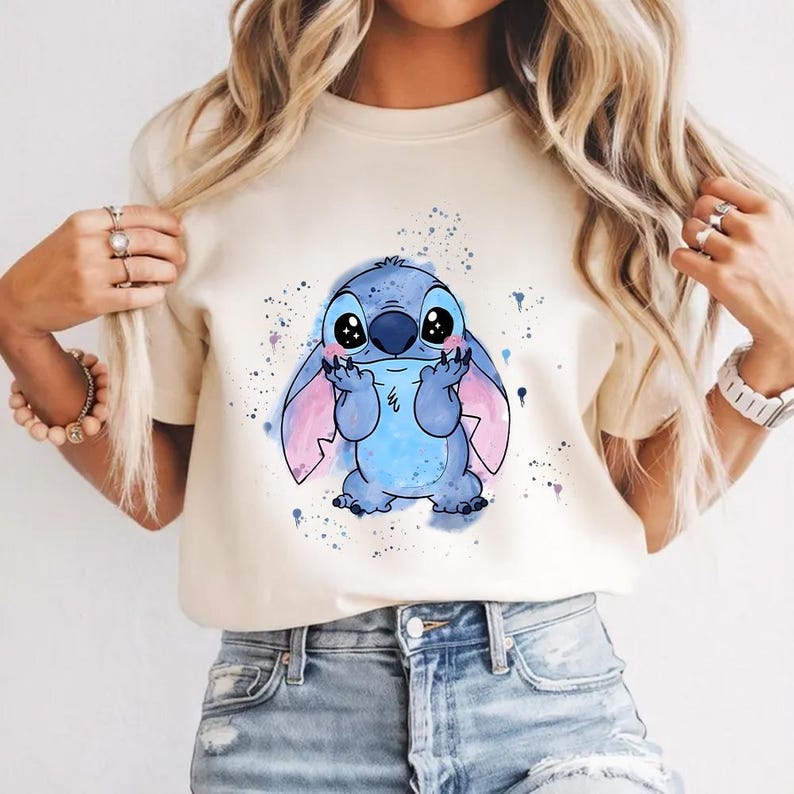 Puede incluir: Camiseta color crema con un personaje de dibujos animados azul con ojos grandes y orejas rosas. El personaje est&aacute; rodeado de salpicaduras de pintura azules y moradas. Es una prenda informal para el d&iacute;a a d&iacute;a.