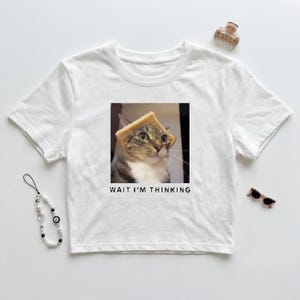 May include: White t-shirt featuring a graphic of a cat wearing toast on its head. The image includes the text "WAIT I'M THINKING" in a black font. The shirt has a classic crew neck design.