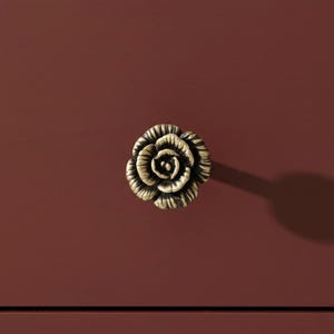 May include: A close-up of a rose-shaped cabinet knob with a bronze finish. The knob is centered on a dark red-brown surface. The rose design has detailed petals and a vintage aesthetic, suitable for furniture hardware.