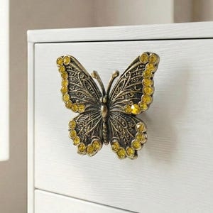 May include: A decorative butterfly-shaped cabinet knob. The knob is made of bronze-colored metal and features intricate detailing on the wings. Yellow gemstones are set around the edges of the wings. The knob is mounted on a white cabinet.