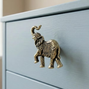 May include: A brass elephant-shaped drawer pull is mounted on a light blue dresser. The elephant has detailed features, including a raised trunk and decorative saddle. The drawer pull adds a unique, decorative touch to furniture.