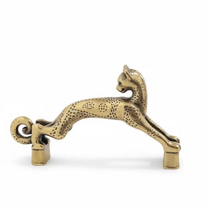 May include: A brass-colored, decorative door knocker in the shape of a stylized leopard. The leopard is arched, with its head and tail raised. The body is detailed with small, circular patterns. The knocker is mounted on two rectangular bases.