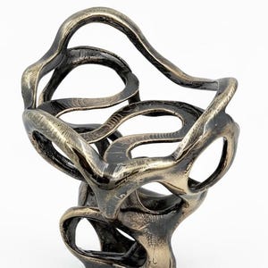 May include: Abstract bronze sculpture with a swirling, open design. The sculpture features interconnected loops and curves, creating an organic, flowing form. The metallic finish has a mix of bronze and dark tones, adding depth and visual interest.