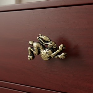 May include: A bronze-colored, leaping rabbit drawer pull mounted on a dark red-brown wooden surface. The rabbit is highly detailed, with its ears back and paws extended. The pull adds a whimsical touch to furniture.