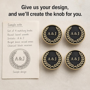 May include: Four round knobs with a charcoal black center and bright brass laurel wreath border. Each knob features the initials A & J in brass. A paper note reads: Give us your design, and we'll create the knob for you. Sample note: Set of 4 matching knobs, Round laurel wreath, Initials: A & J, Bright brass raised areas, Charcoal black recesses, Sample design.