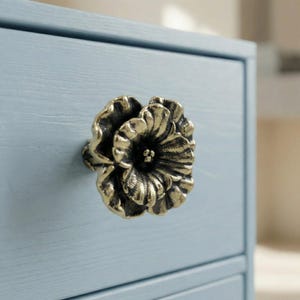 Könnte beinhalten: Ein bronzefarbener, blütenförmiger Schubladengriff ist an einem hellblauen Schrank befestigt. Der Schubladengriff hat detaillierte Blütenblätter und eine dunkle Mitte. Der Schrank hat eine lackierte Oberfläche und sichtbare Holzmaserung.