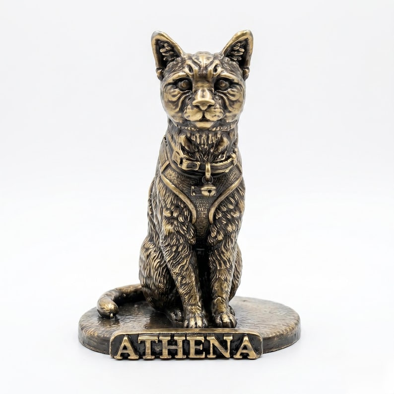 May include: A bronze-toned cat figurine sitting on a circular base. The cat wears a harness with a small bell attached to the collar. The name ATHENA is embossed in capital letters on the front of the base. The sculpture has a textured finish.