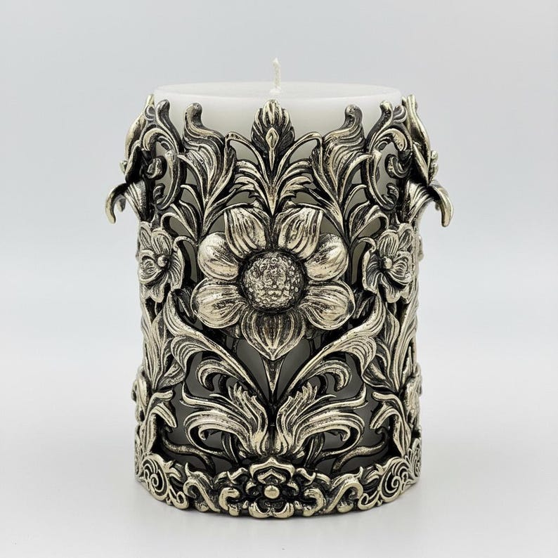 Pu&ograve; includere: Una candela a colonna bianca all'interno di un portacandele in metallo argentato ornato. Il portacandele presenta un design floreale con foglie e fiori dettagliati. La candela ha un'unica miccia.