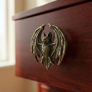 May include: A bronze-colored bat-shaped cabinet pull with outstretched wings, mounted on a dark wood drawer. The bat has detailed features, including a textured body and wing design. The hardware adds a gothic or fantasy theme to furniture.
