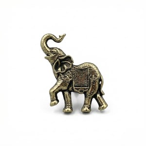 May include: A detailed bronze-colored elephant figurine with its trunk raised. The elephant has intricate carvings on its body and a decorative saddle. The figurine is set against a plain white background, highlighting its ornate details.