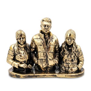 May include: A metallic gold-toned sculpture depicting three figures standing side-by-side on a flat base. The central figure wears a puffer jacket, while the two flanking figures wear beanies and puffer jackets with long hair visible.