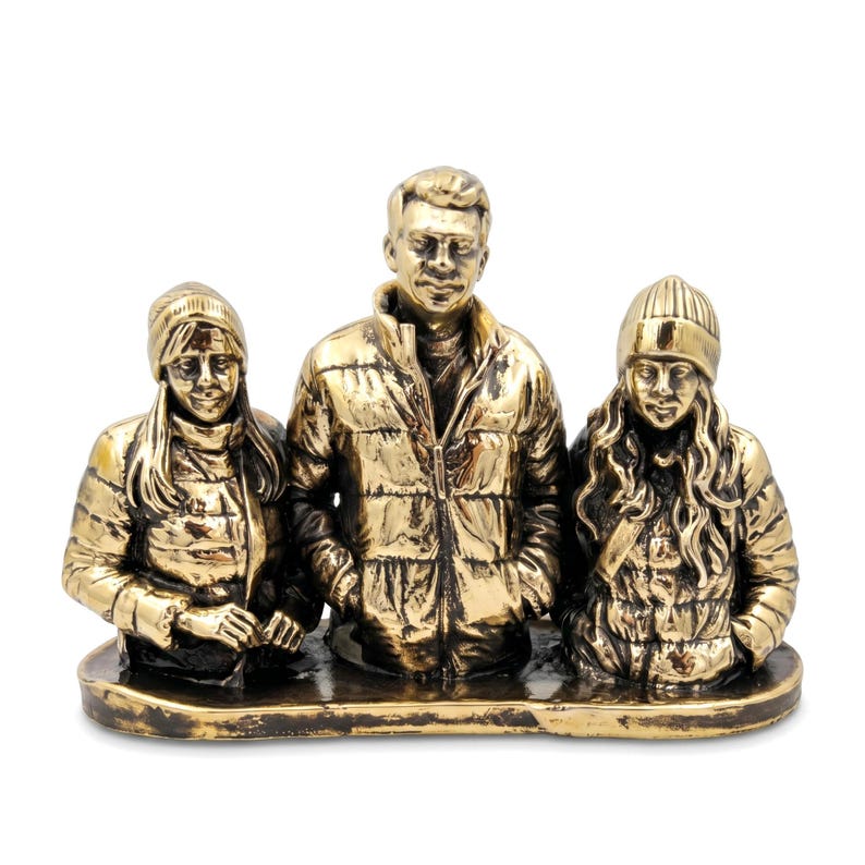 May include: A metallic gold-toned sculpture depicting three figures standing side-by-side on a flat base. The central figure wears a puffer jacket, while the two flanking figures wear beanies and puffer jackets with long hair visible.