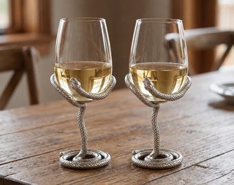 Könnte beinhalten: Zwei Weingläser aus klarem Glas mit silberfarbenen Metallstielen und Sockeln in Form von gewundenen Schlangen. Die Schlangenkörper umschlingen den unteren Teil jedes Glases, wobei die Köpfe direkt an der Glasoberfläche anliegen.