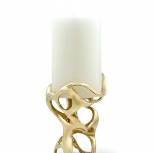 May include: A gold-colored candle holder with an abstract, organic design. The holder has a swirling, openwork pattern that supports a large, white pillar candle. The candle holder is made of metal and has a smooth, reflective surface.