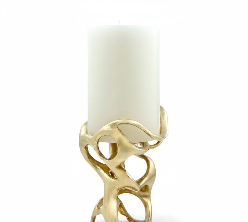 May include: A gold-colored candle holder with an abstract, organic design. The holder has a swirling, openwork pattern that supports a large, white pillar candle. The candle holder is made of metal and has a smooth, reflective surface.