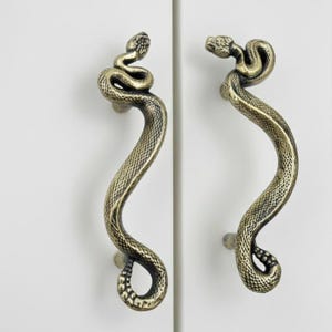 May include: Two antique brass cabinet handles in the shape of snakes. The handles are detailed with scales and a coiled head. The handles are mounted on a white cabinet door.
