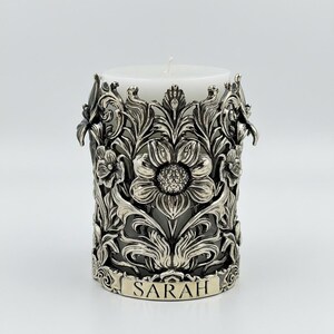 Pu&ograve; includere: Una candela a colonna bianca all'interno di un portacandele floreale color argento. Il portacandele presenta un design intricato di fiori e foglie, con il nome "SARAH" inciso sulla base. La candela &egrave; non profumata.