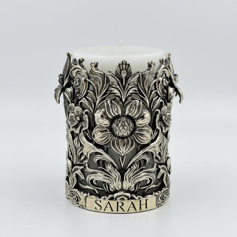 Pu&ograve; includere: Una candela a colonna bianca all'interno di un portacandele floreale color argento. Il portacandele presenta un design intricato di fiori e foglie, con il nome "SARAH" inciso sulla base. La candela &egrave; non profumata.