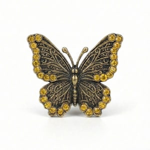 May include: A detailed butterfly-shaped brooch with a bronze finish. The wings are adorned with yellow gemstones and intricate filigree patterns. The brooch is a decorative accessory.