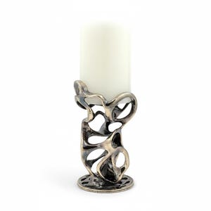 May include: A decorative candle holder with an abstract, openwork design in a metallic finish. The holder supports a white pillar candle. The base is round with a patterned edge. The overall aesthetic is modern and artistic.