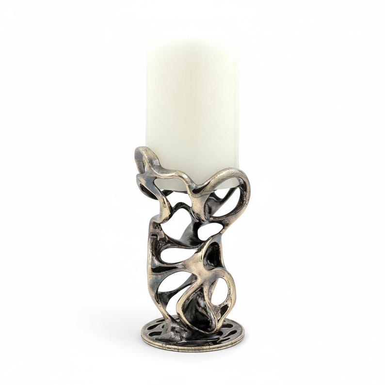May include: A decorative candle holder with an abstract, openwork design in a metallic finish. The holder supports a white pillar candle. The base is round with a patterned edge. The overall aesthetic is modern and artistic.