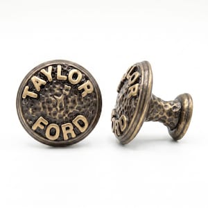 Peut inclure: Deux boutons de placard en métal de couleur bronze avec une texture martelée. La face avant de chaque bouton présente le texte en relief "TAYLOR FORD" dans une police sans empattement. Les boutons ont une base cylindrique et une plaque.