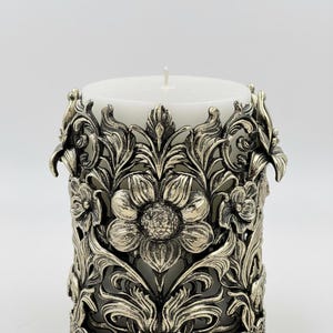 Pu&ograve; includere: Una candela a colonna bianca &egrave; all'interno di un manicotto decorativo in metallo color argento. Il manicotto presenta un intricato design floreale con foglie e fiori dettagliati. La candela ha un'unica miccia bianca.