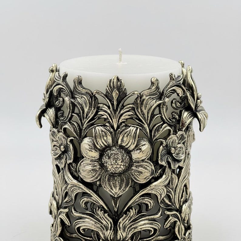 Pu&ograve; includere: Una candela a colonna bianca &egrave; all'interno di un manicotto decorativo in metallo color argento. Il manicotto presenta un intricato design floreale con foglie e fiori dettagliati. La candela ha un'unica miccia bianca.