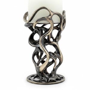 May include: A decorative candle holder with a unique, openwork design. The metal holder has a dark bronze finish with lighter bronze accents. A white candle sits in the center of the holder. The base is round with a decorative pattern.