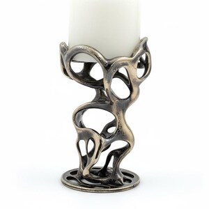 May include: An abstract bronze-colored candle holder with a white candle. The holder has an open, organic design with a circular base and a top that cradles the candle. The candle is a thick, cylindrical shape.